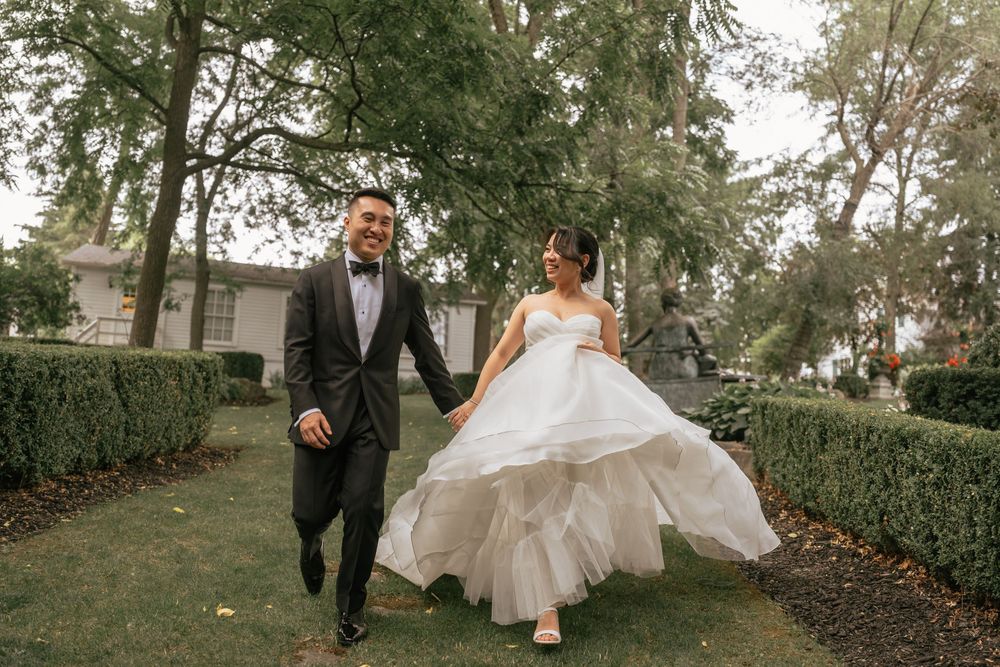 A couple in wedding attire walks hand-in-hand through a lush garden, the bride lifting her white gown.