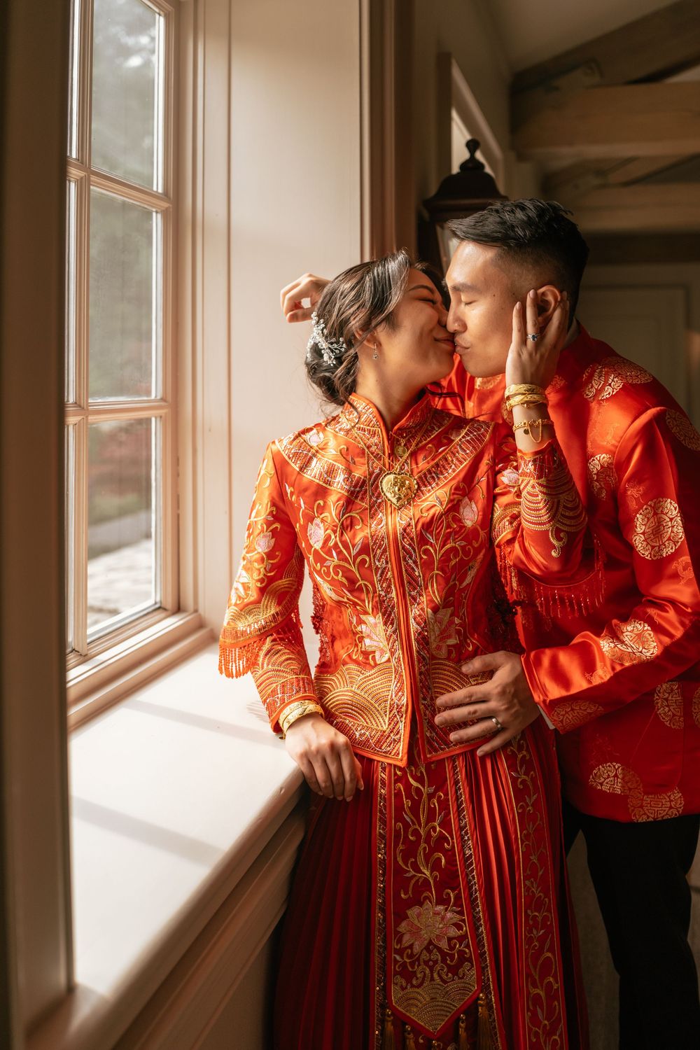 Asian couple shares a romantic kiss wearing traditional red Chinese wedding attire beside a sunlit window.