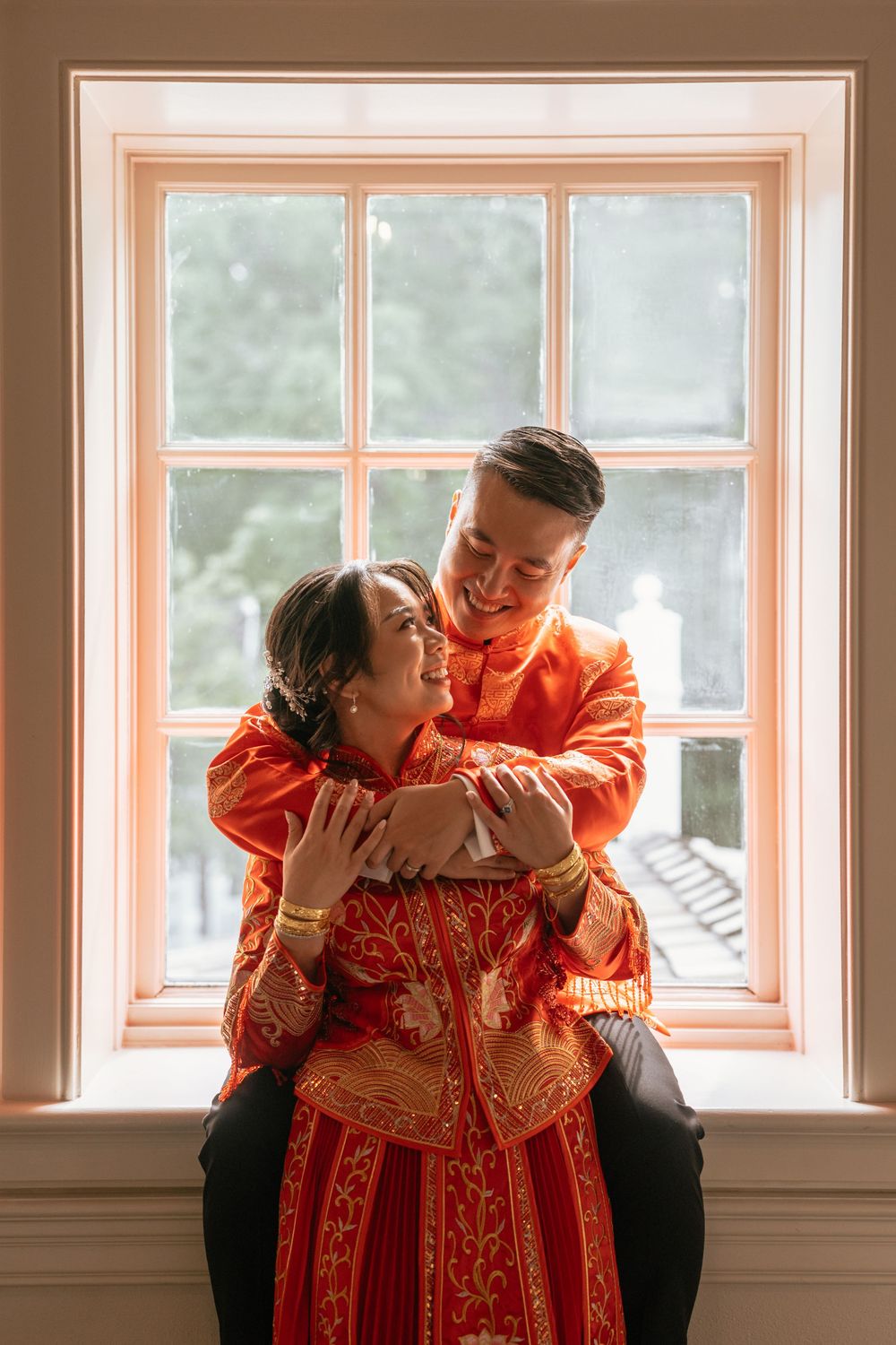 Couple in traditional red Chinese wedding attire embracing by a window, smiling at each other.
