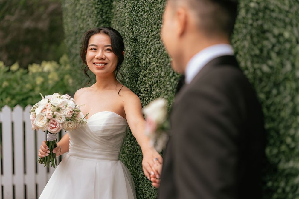 Radiant Asian bride in strapless white gown holds bouquet smiling at groom before a lush green hedge wall.