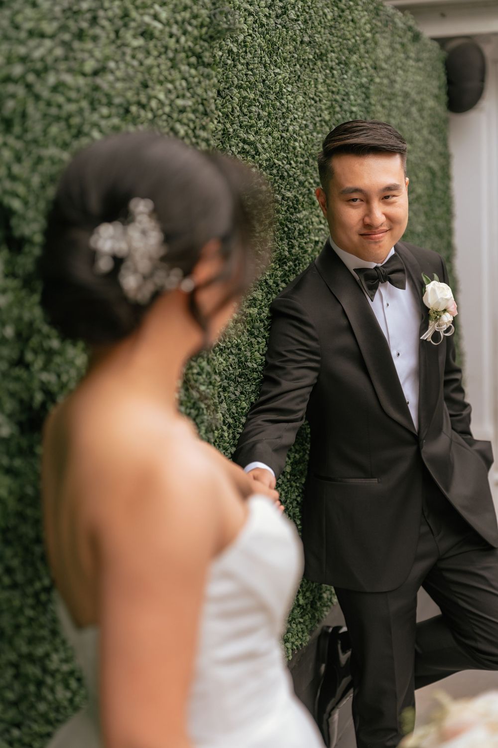 Groom in black tuxedo smiling at bride while leaning against a lush green hedge wall.