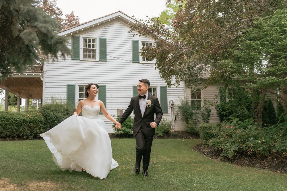 Bride and groom walking hand in hand on green lawn in front of white colonial house with green shutters.