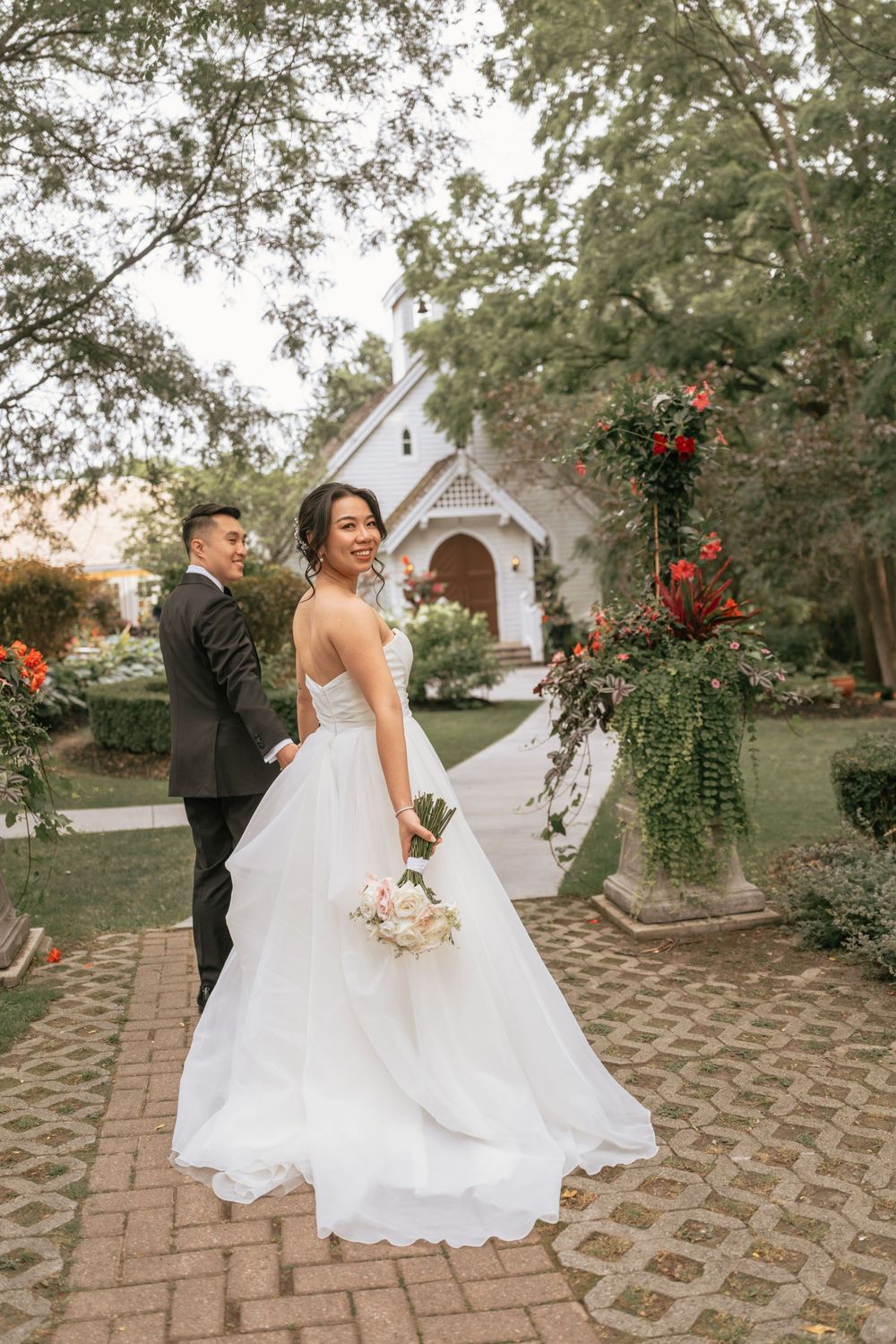 Bride in white ball gown holds bouquet with groom in dark suit outside a charming chapel surrounded by lush greenery.