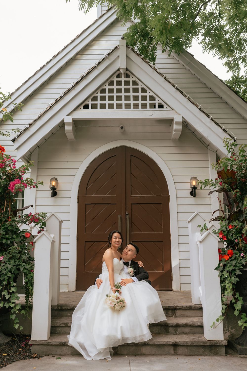 Bride and groom sitting on steps of a charming white chapel surrounded by colorful flowers.