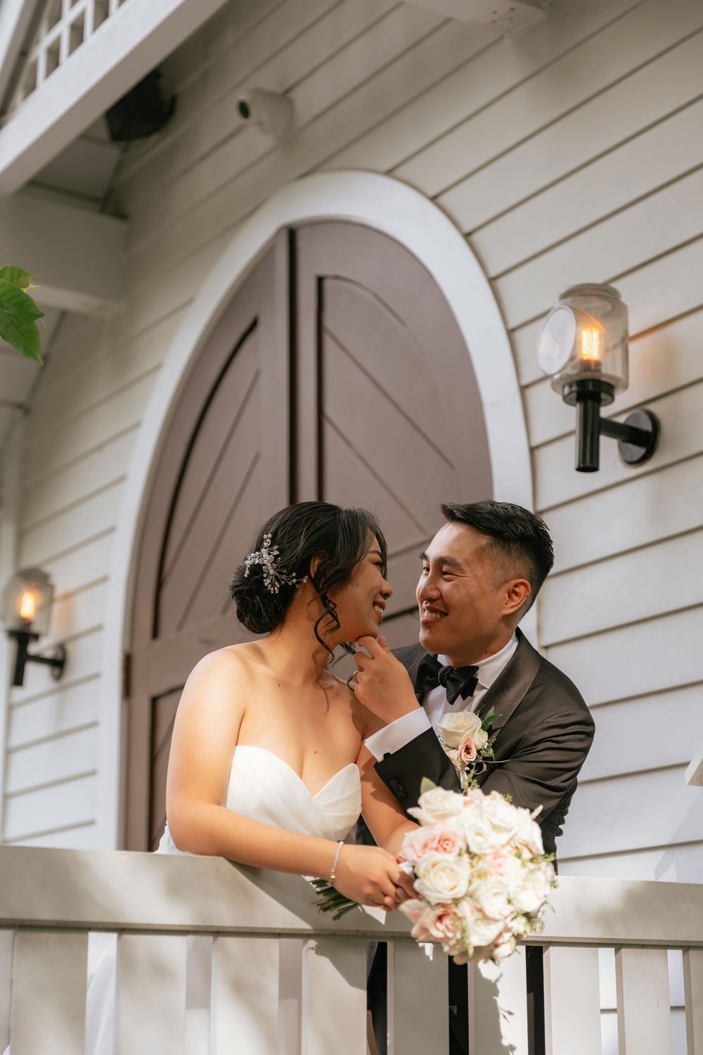 Bride and groom sharing a romantic moment on a white chapel balcony, holding a bouquet of roses.