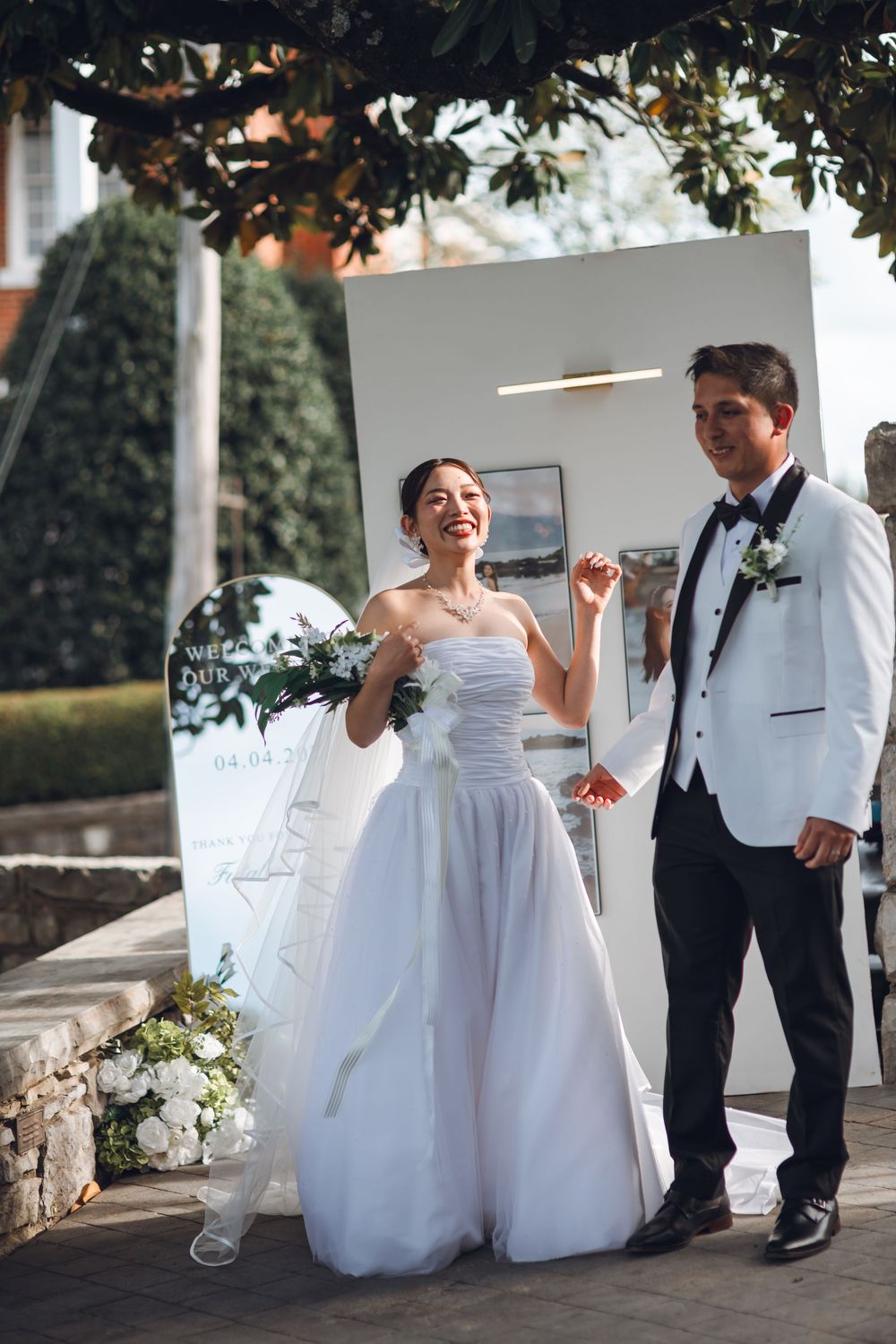 Happy bride in white strapless gown and groom in white tuxedo posing outdoors at elegant wedding ceremony.