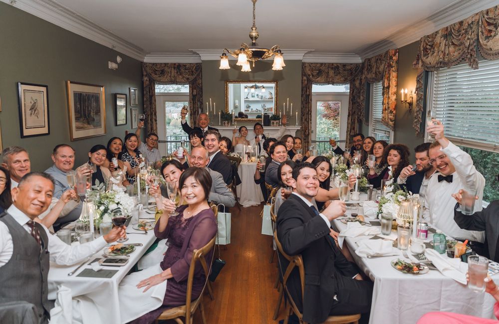 Wedding reception guests raising glasses for a toast in an elegant dining room with white tablecloths and floral centerpieces.
