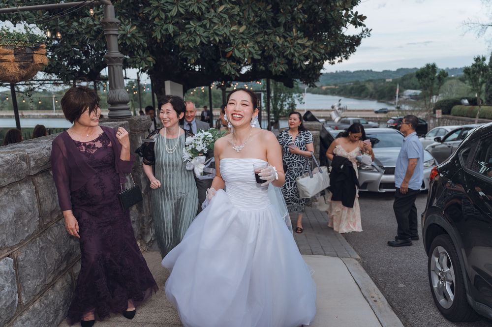 Smiling bride in white wedding gown walks with guests outdoors near a stone wall and parked cars.
