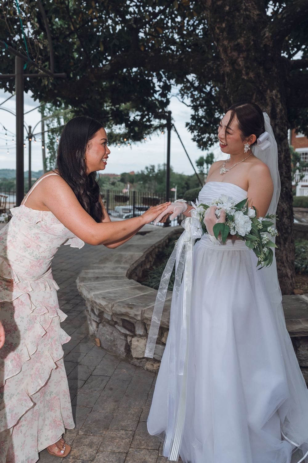 A bridesmaid adjusts the bride's white gown as she holds a floral bouquet at an outdoor wedding venue.