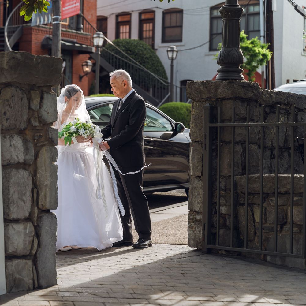 Bride in white gown and veil stands with older man in black suit near iron gate and dark car on sunny day.