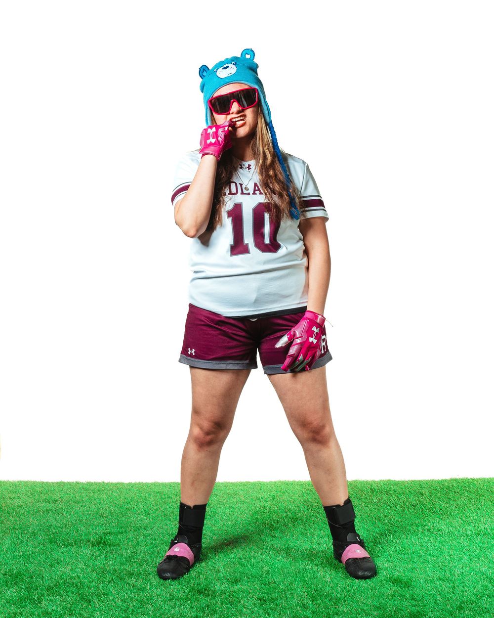 Female athlete in jersey number 10, maroon shorts, pink gloves, and a blue bear hat posing on turf.