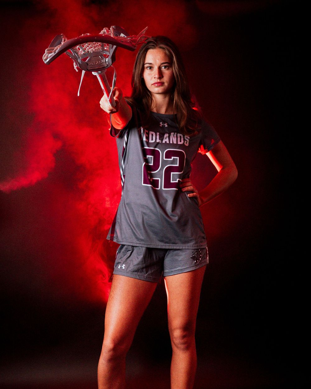 Female lacrosse player #22 in Redlands uniform poses with stick amid red smoke on dark background.