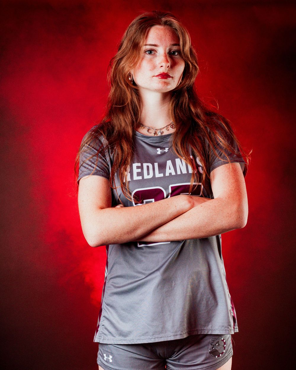 Young athlete in Redlands jersey poses confidently with arms crossed against dramatic red smoky background.