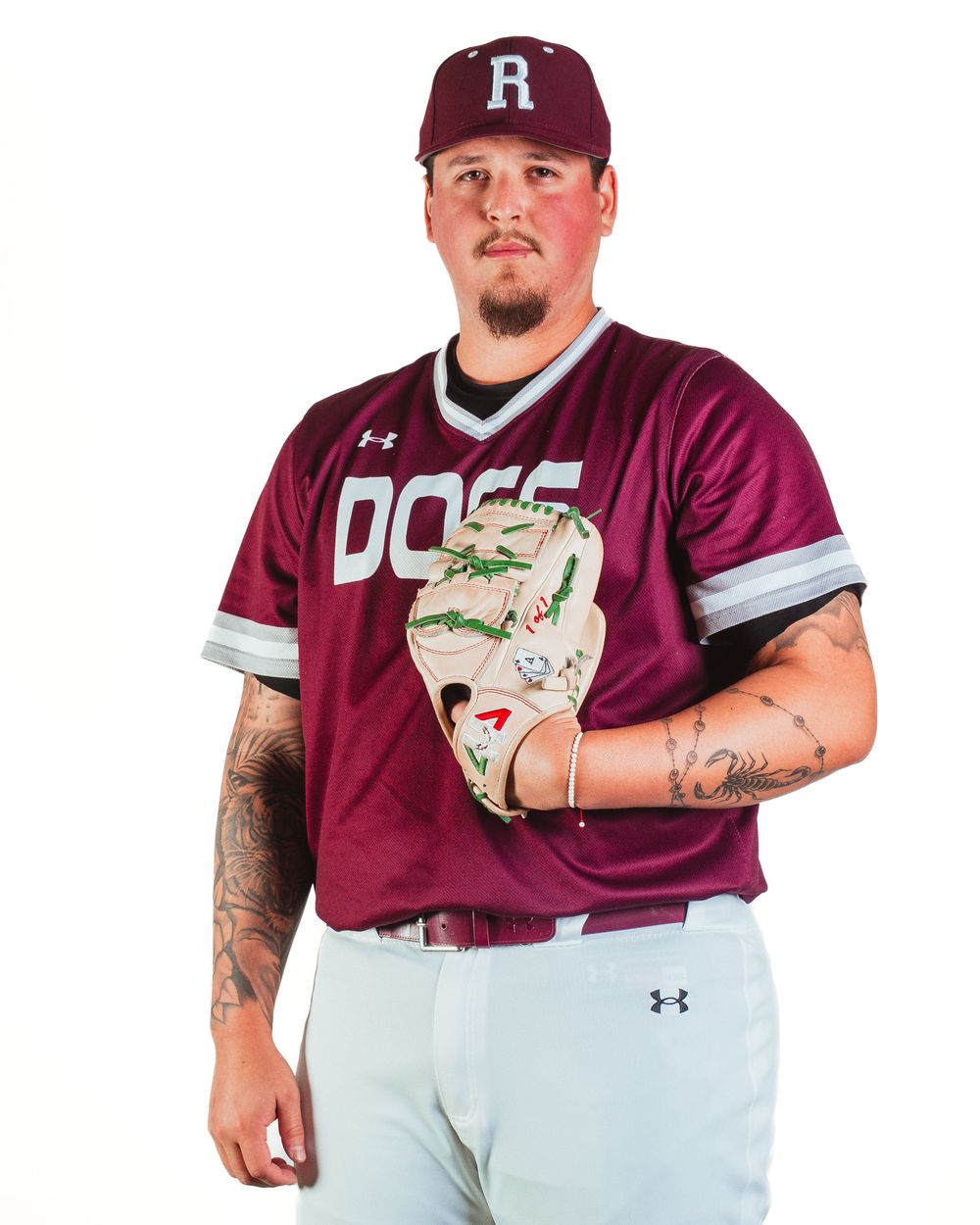 Baseball player in maroon Dogs jersey and cap holding a glove, wearing Under Armour uniform with tattooed arms.
