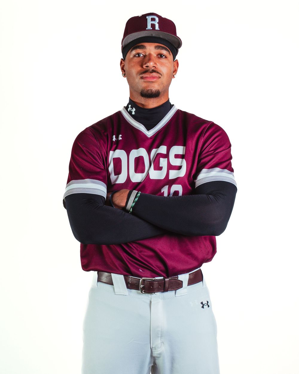 Baseball player in maroon DOGS jersey and cap with arms crossed, wearing Under Armour gear and arm sleeves.