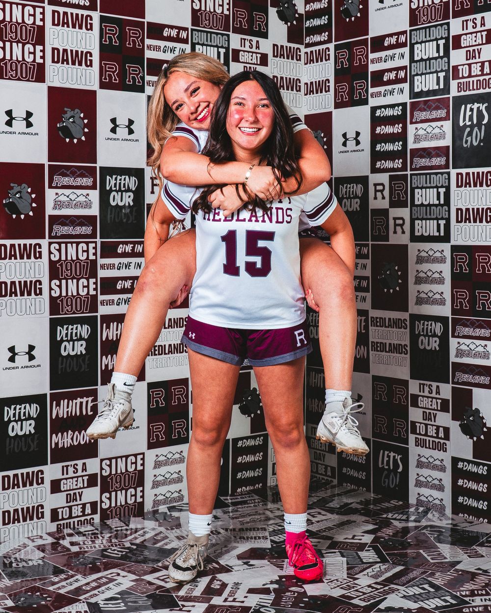 Two female athletes in Redlands uniforms pose playfully, one giving the other a piggyback ride in front of a maroon photo backdrop.