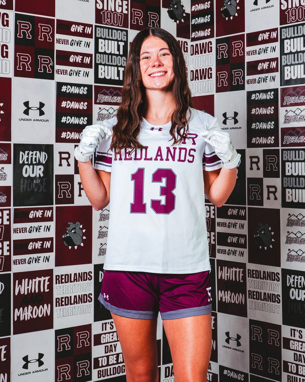 Female athlete wearing Redlands jersey number 13 poses in front of school spirit backdrop.