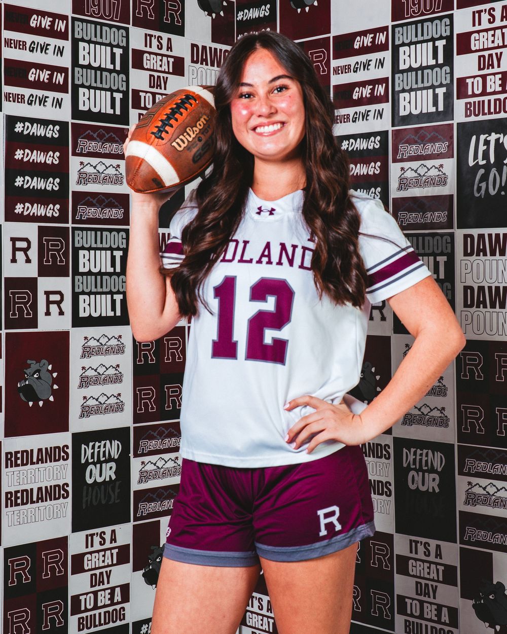 Female athlete in Redlands jersey number 12 holds football, posing confidently in front of Bulldogs branded backdrop.