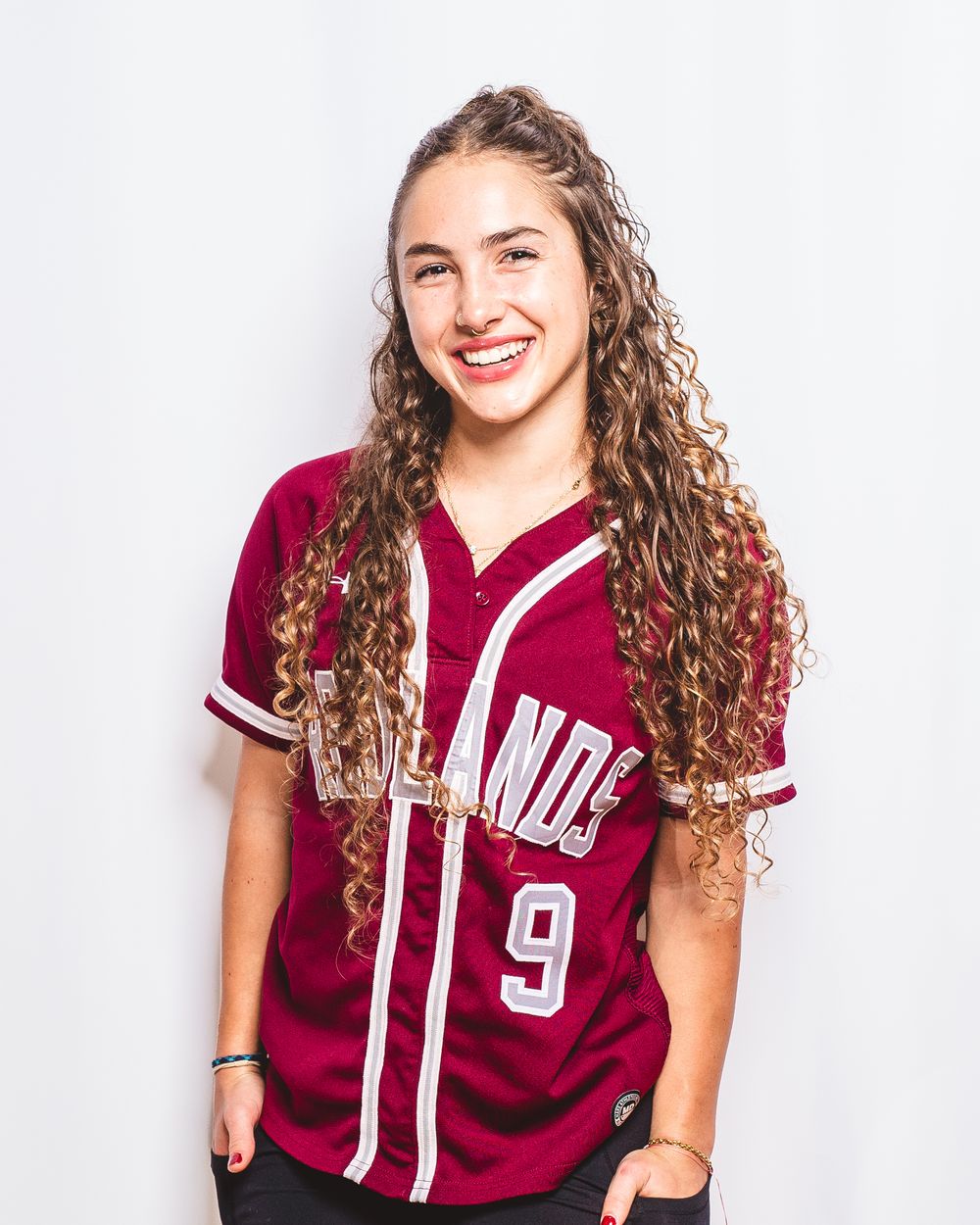 Smiling young woman with curly hair wearing a maroon baseball jersey number 9 posing against white background.