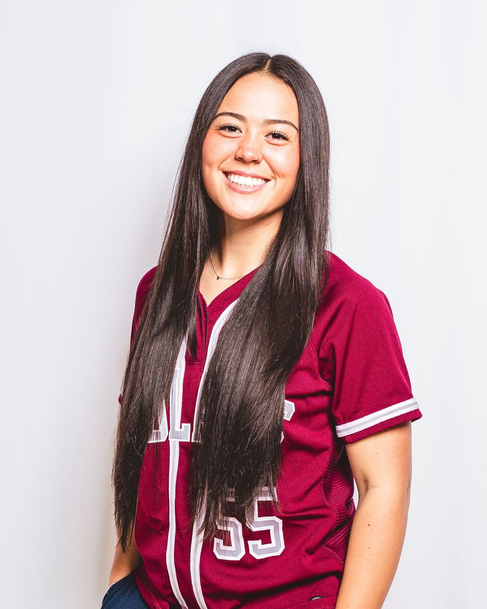Young woman in maroon baseball jersey number 55 smiling, with long dark hair, against white background.