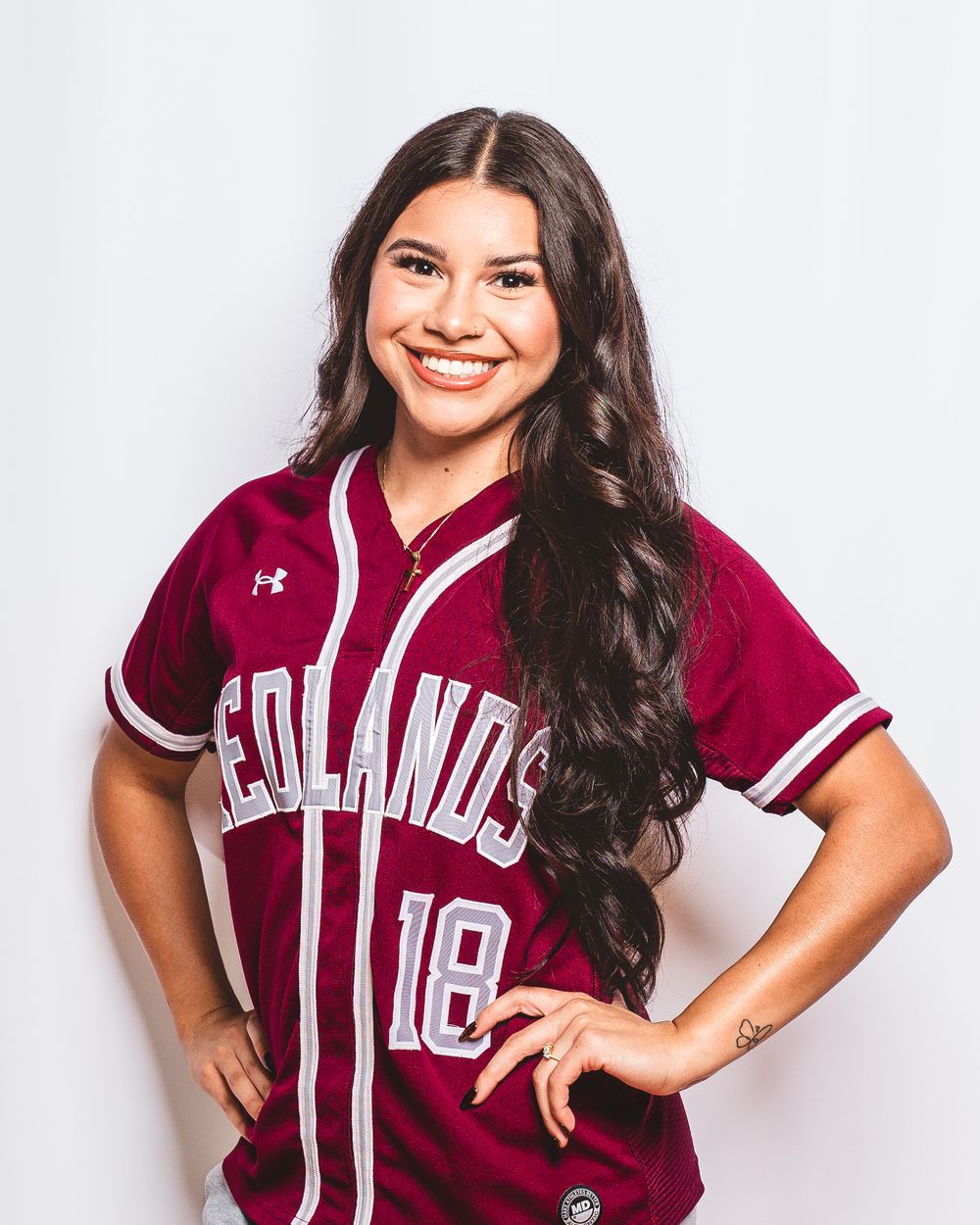 Young woman in a maroon Redlands #18 Under Armour softball jersey smiling confidently with hands on hips.