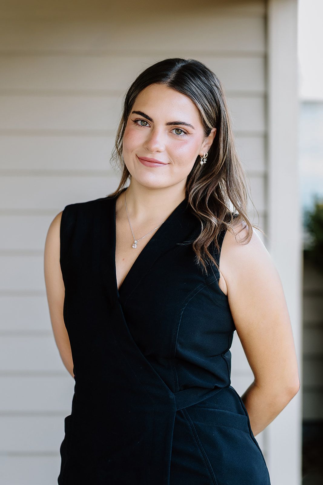 Young woman in black sleeveless dress smiling outdoors, wearing delicate jewelry against neutral background.