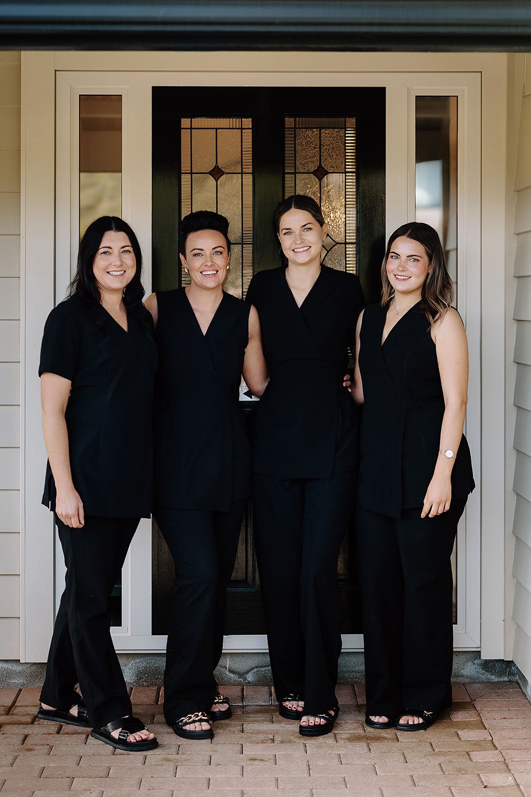 Four women in matching black uniforms smile together in front of an elegant door, representing a professional spa team.