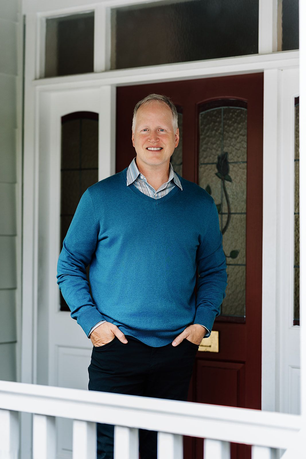 Smiling man in blue sweater standing on porch in front of red door with decorative glass panels.