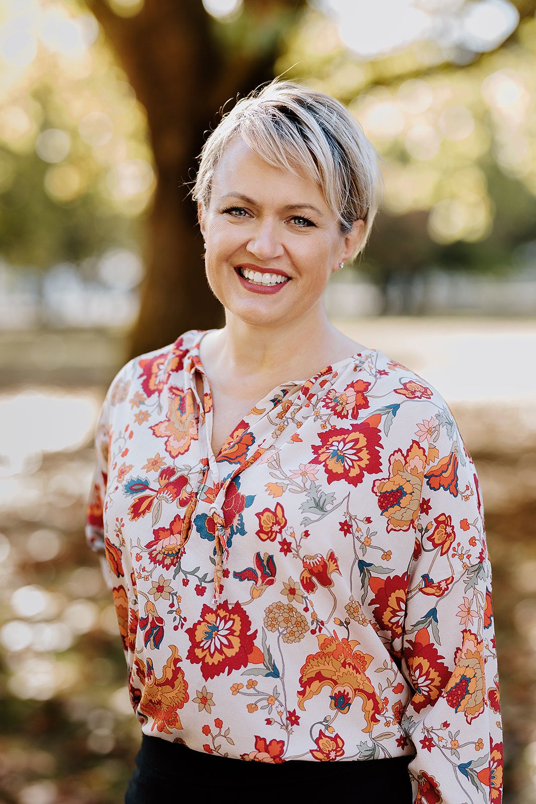 Smiling woman with short blonde hair wearing a floral blouse, posing outdoors in a sunlit park.