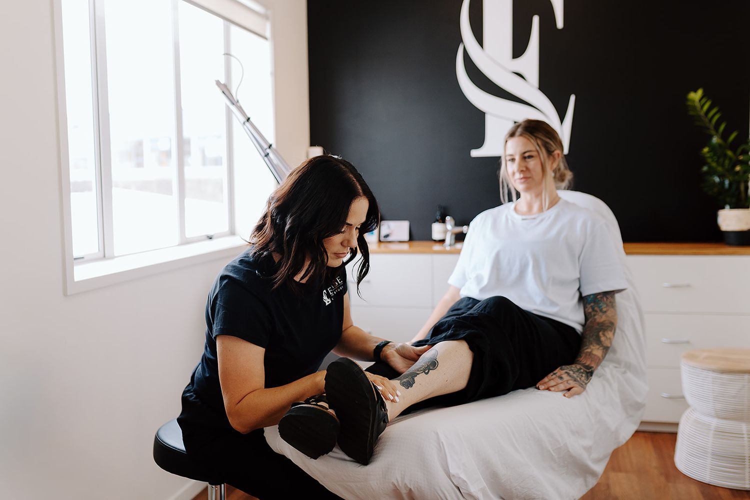 Tattoo removal specialist treating a client's ankle tattoo in a modern studio with black accent wall.