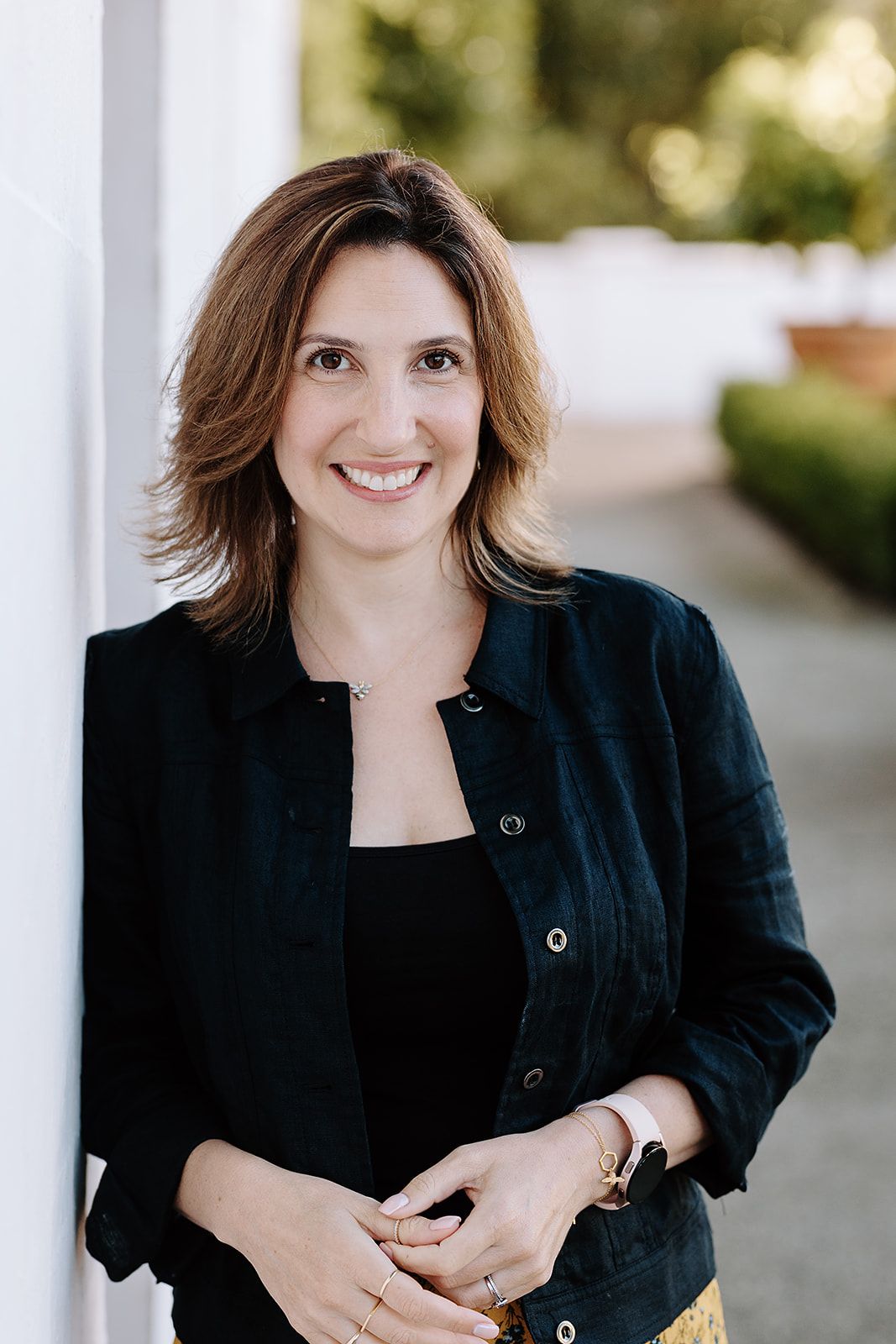 Smiling woman with brown hair in black jacket leaning against white column outdoors.