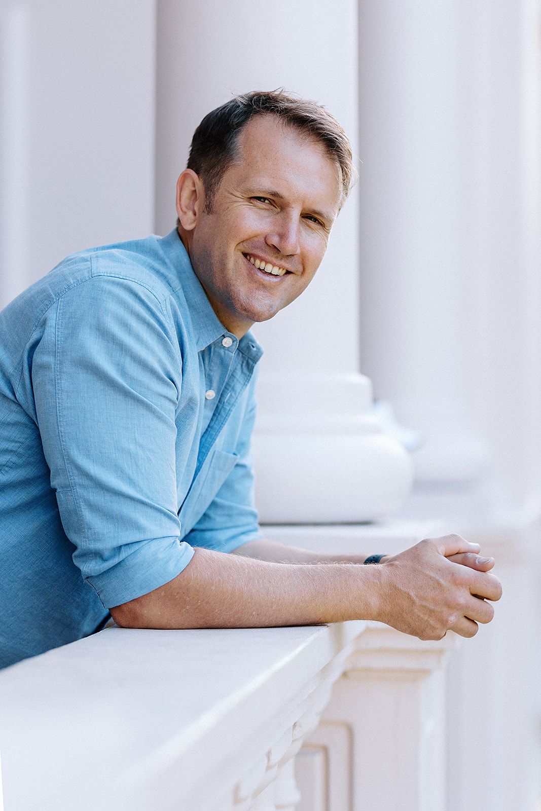 Smiling middle-aged man in blue shirt leaning on white balustrade with classical columns in background.