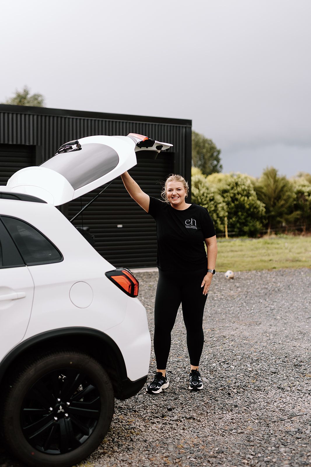 Woman in black activewear opening the trunk of a white SUV on a gravel driveway outside a black building.