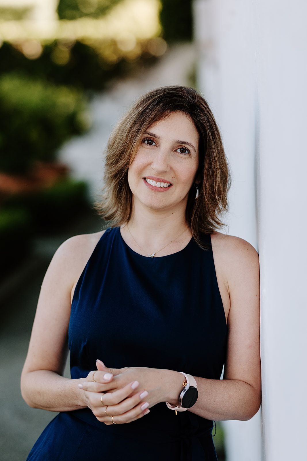 Smiling woman with brown hair in a navy sleeveless dress, leaning against a white wall outdoors.