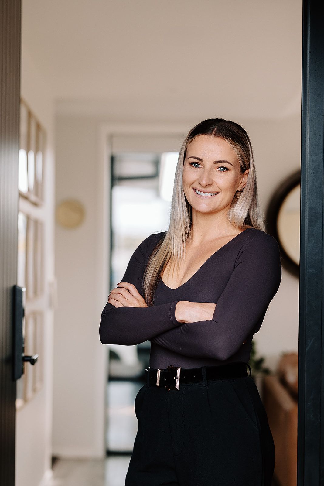 Confident smiling woman in black outfit standing with arms crossed in a modern home doorway.