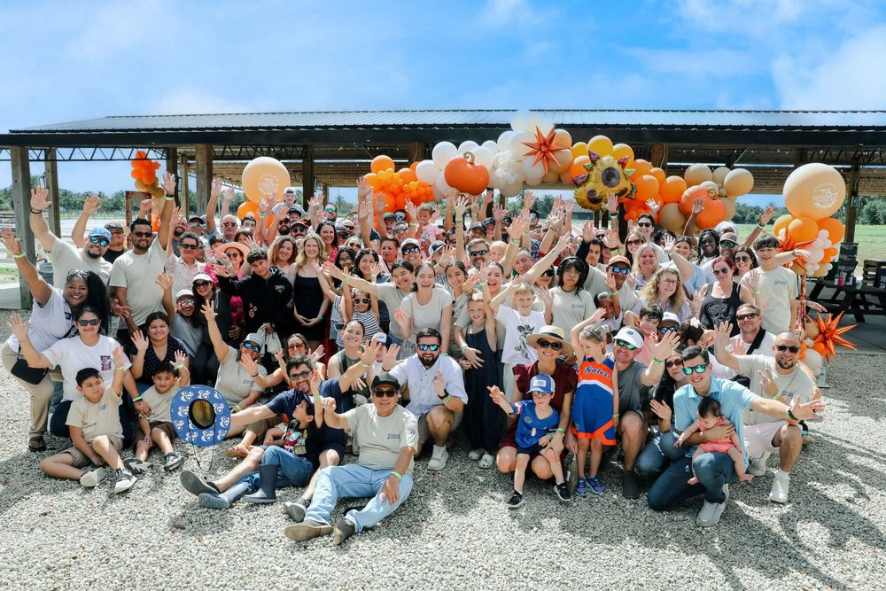Large group of people celebrating outdoors with orange and white balloons and floral decorations at a festive event.