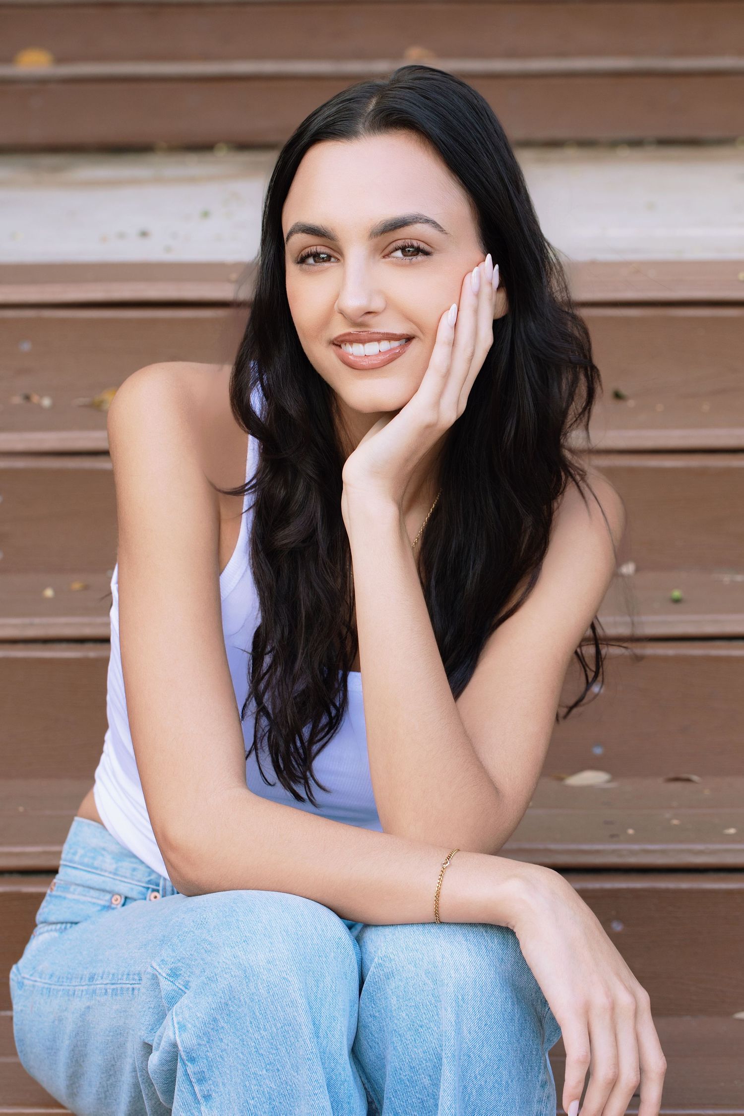 Young woman with long dark hair smiling, sitting on wooden steps in a white tank top and blue jeans.
