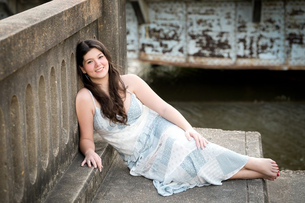 Young woman in floral dress sitting on stone steps near water, smiling outdoors.