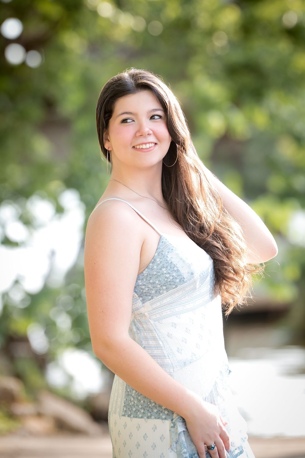 Young woman in light blue floral sundress smiling outdoors with lush green trees in soft bokeh background.