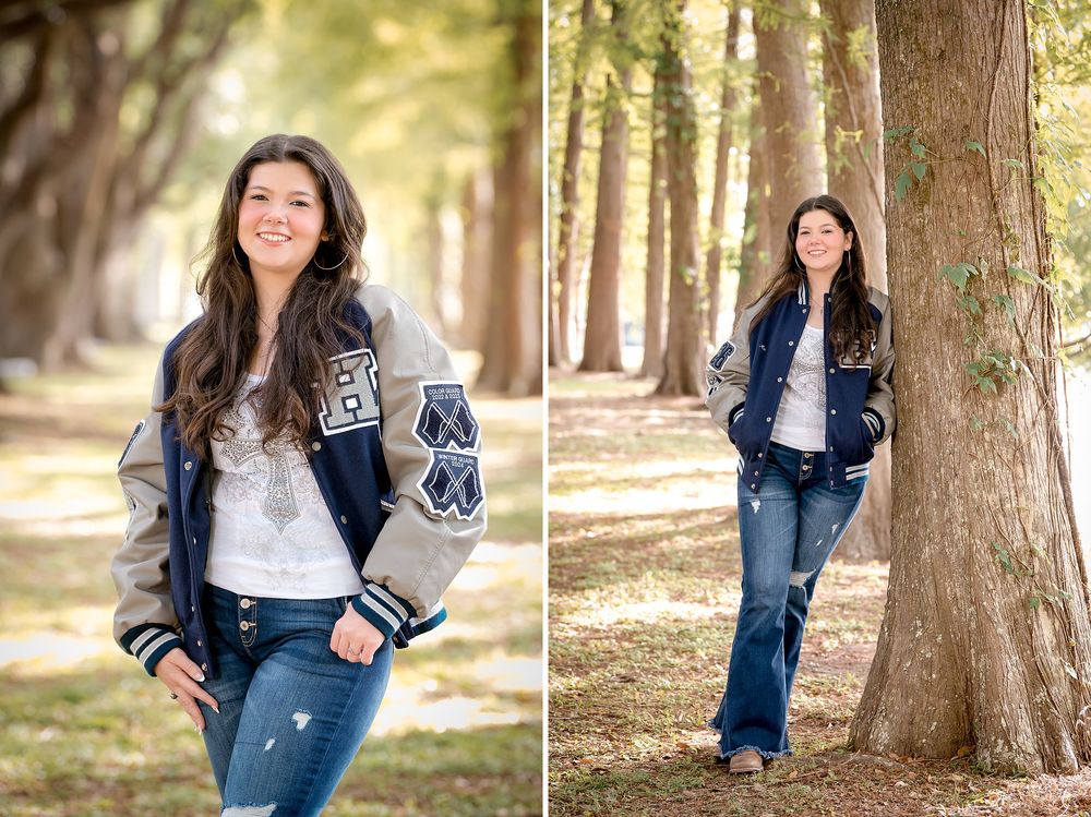 Teen girl in navy letterman jacket and jeans smiling outdoors among trees in a senior portrait photo session.