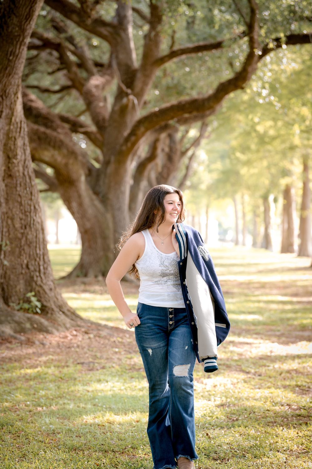Young woman in ripped jeans and white tank top walks through a tree-lined park, smiling and carrying a navy jacket.