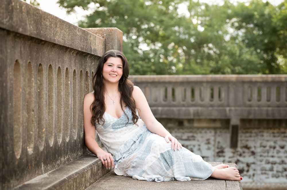 Young woman in flowy blue dress sitting on stone steps outdoors, smiling warmly in a garden setting.