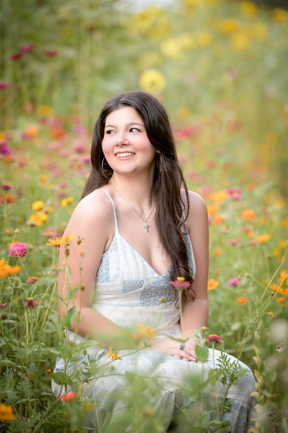 Young woman in white dress sitting in colorful wildflower field, smiling, wearing cross necklace, surrounded by pink and orange blooms.