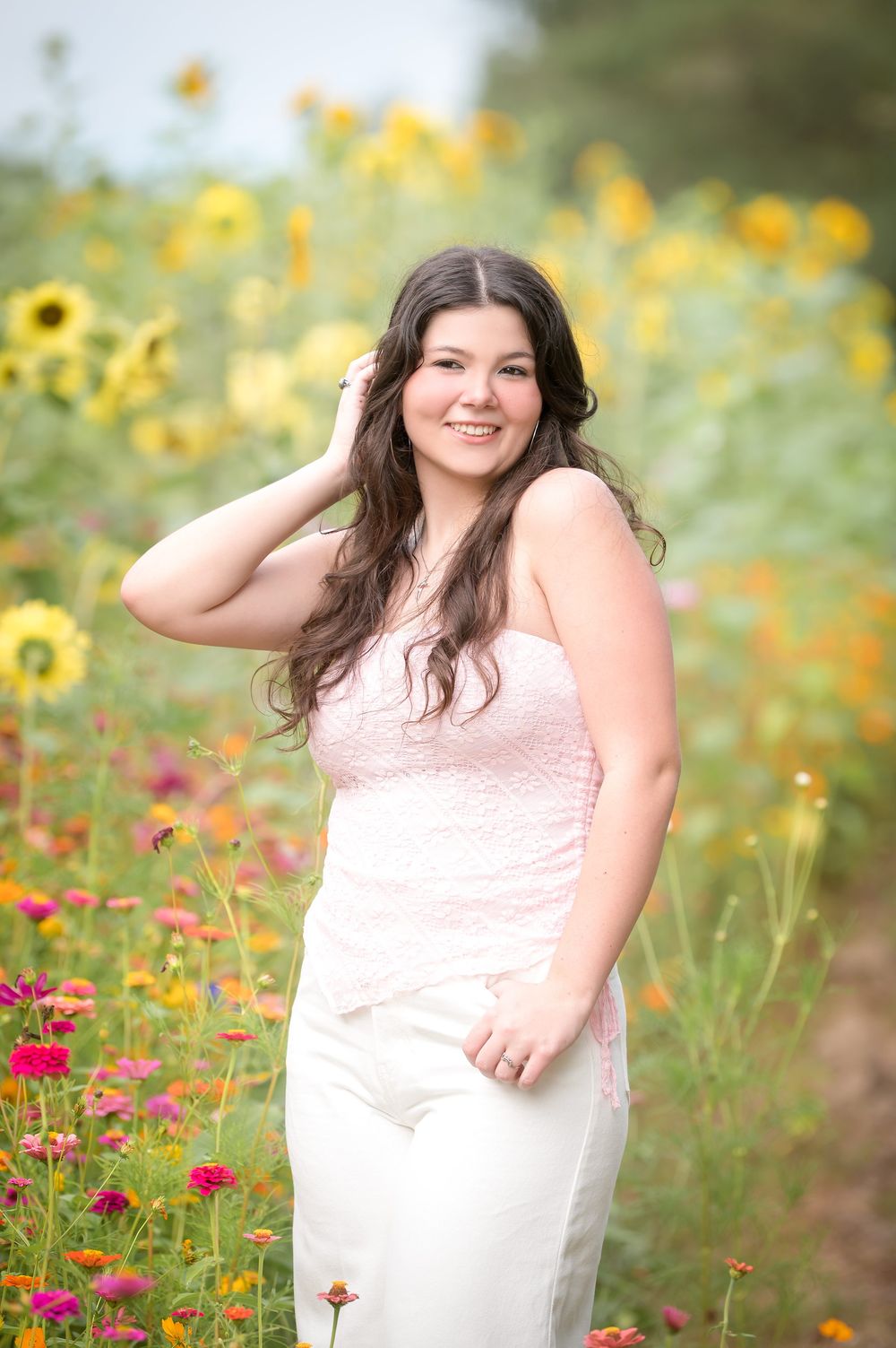 Young woman in pink top and white pants smiling in a vibrant wildflower field with sunflowers.