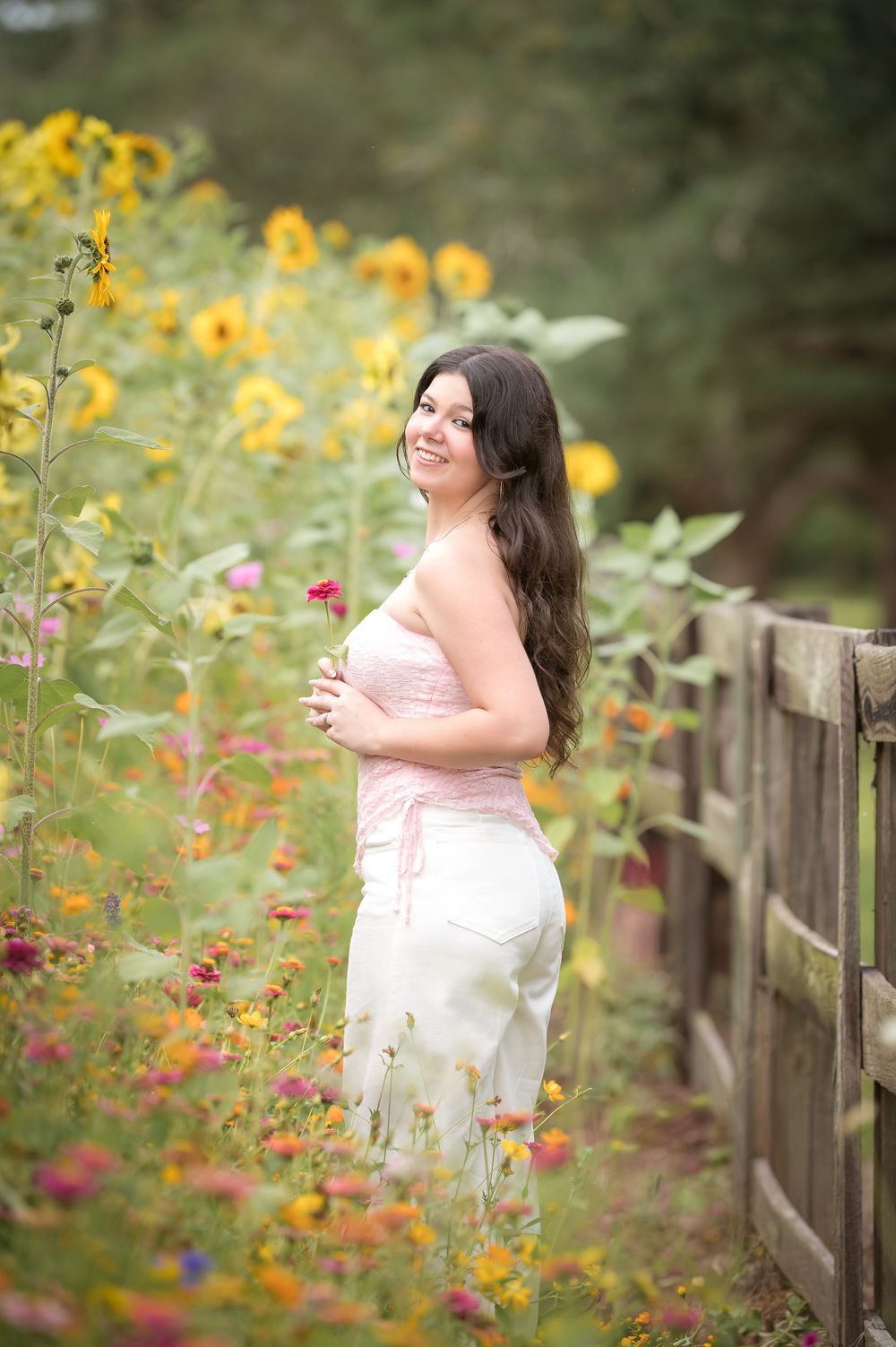 Young woman in pink top and white jeans smiling among colorful wildflowers and sunflowers beside a wooden fence.