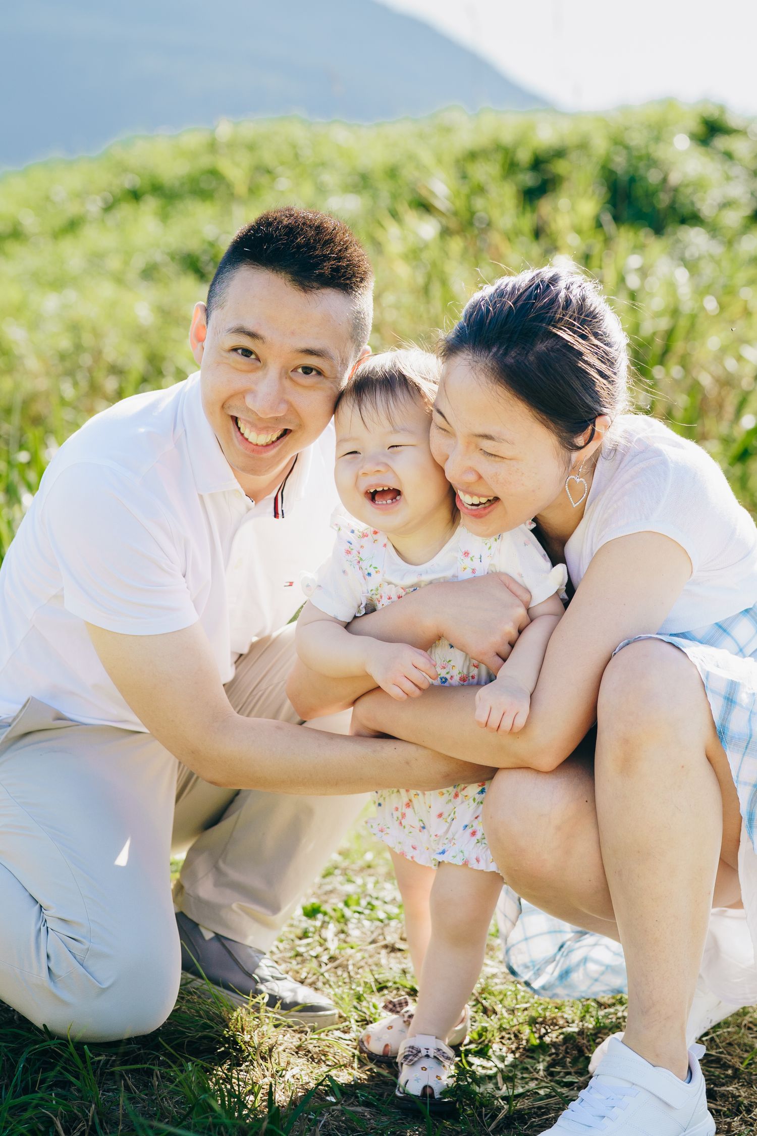 Happy Asian family with smiling parents and laughing baby girl sitting outdoors on green grass with mountains behind.