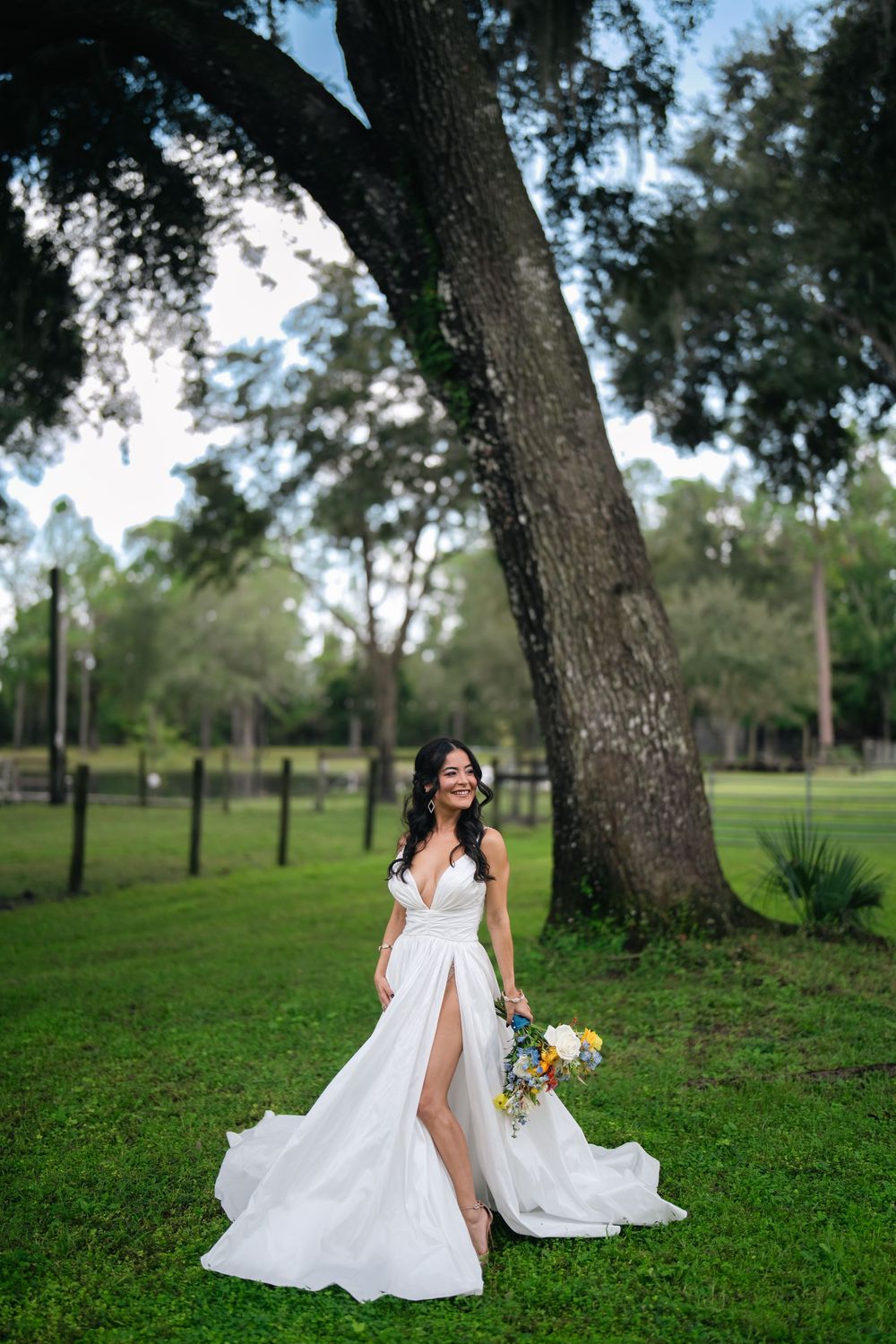 Bride in a white gown with a slit holds a bouquet, smiling outdoors near a large oak tree on green grass.