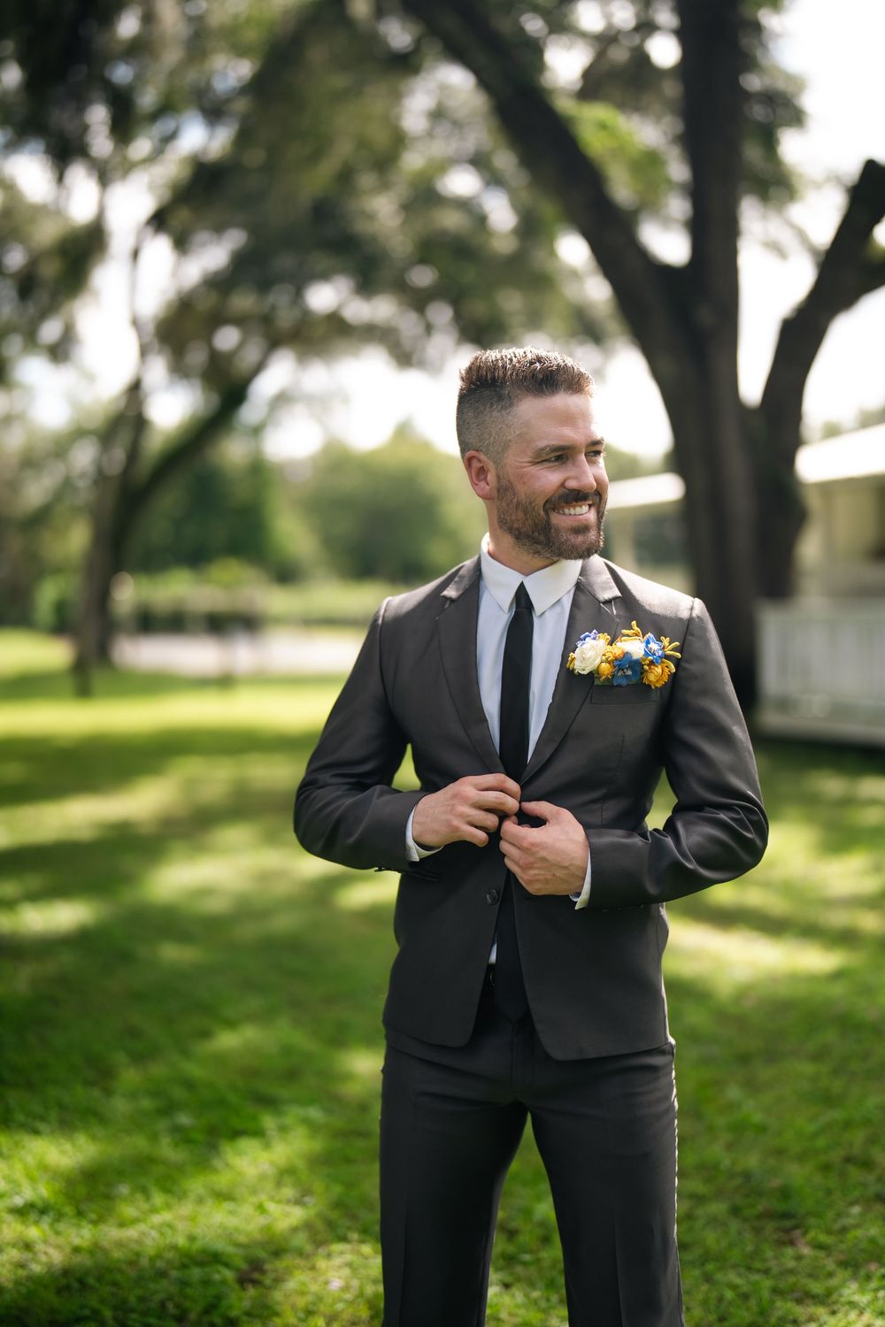 Smiling groom in gray suit with boutonniere buttoning jacket outdoors in lush green park setting.