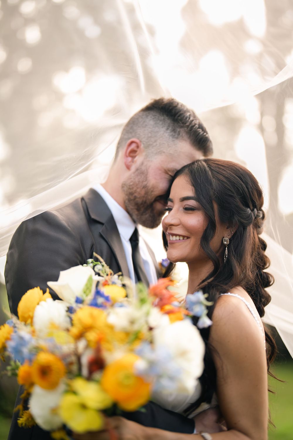 Bride and groom share romantic moment under veil with colorful wedding bouquet of yellow, white, and blue flowers.