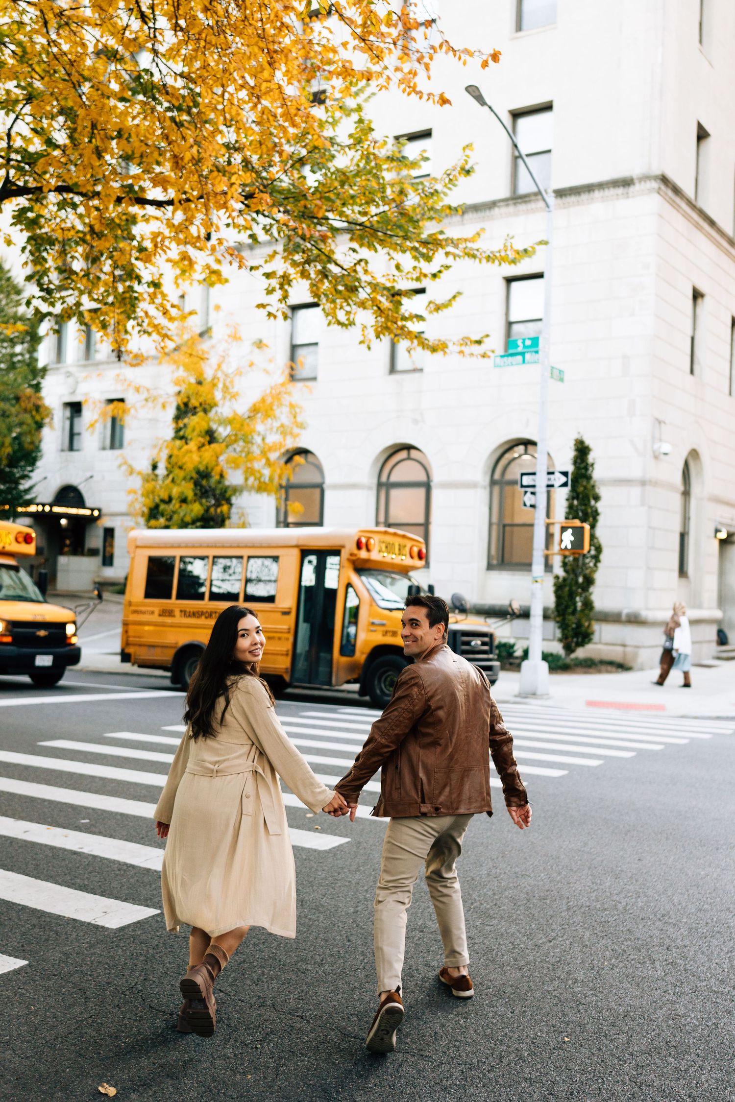 A couple holds hands while crossing a city street with fall foliage and a yellow school bus in view.